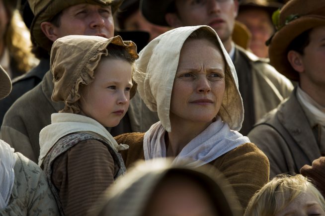 A woman and child in 19th-century clothes stares with determination to the right, amongst a busy crowd of protesters in this still from Peterloo.