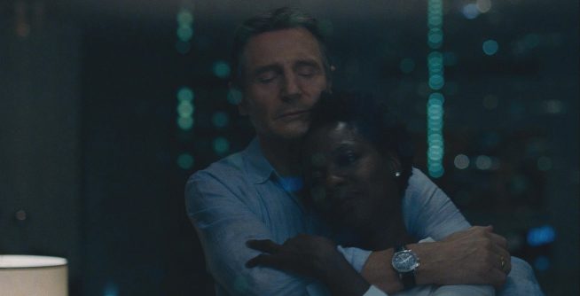 A middle-aged African American woman leans into the affectionate embrace of a middle-aged white man in a light blue shirt in this image from Widows. In the background can be seen blurry, soft green lights.