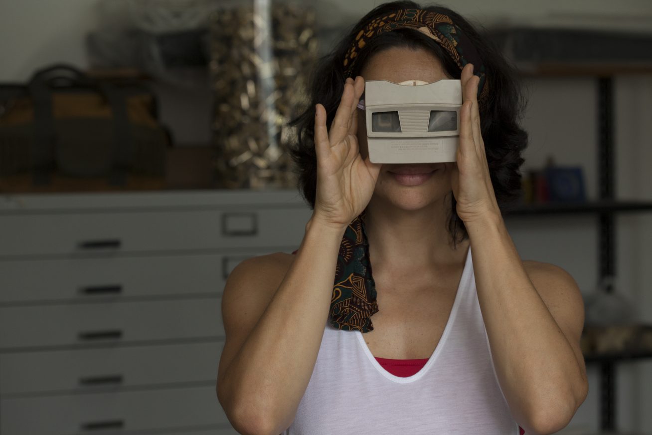 A woman in a cluttered room holds an old slide camera up to her face as she faces the camera in this still from Cinema of Brazil 2018: Female Perspectives.