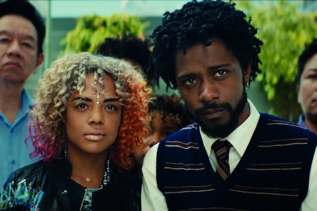 A young African American woman with multi-coloured hair stands next to a young, bearded African American male in a shirt, tie and sleeveless blazer. Both look into the distance beyond the camera in this still from Sorry to Bother You.
