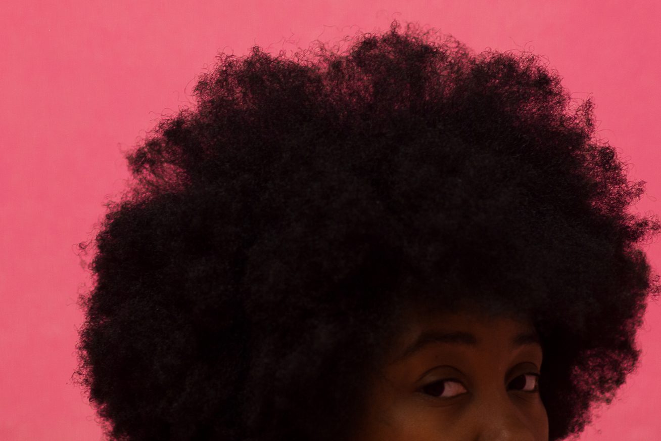 Against a bright, pink background, we see the face of a young black woman, side on, from the nose up. She glances sideways at us, and her large afro fills most of the lower half of this Afrofuturism still from London Short Film Festival: No Woman Is An Island.