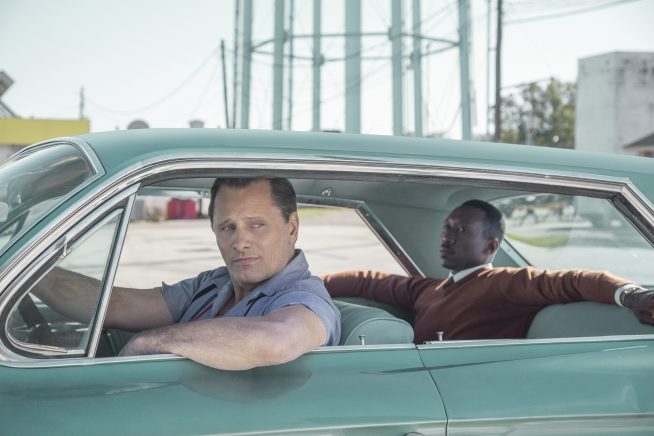 In a deserted looking industrial estate, two men sit in the foreground in a pretty turquoise American Cadillac. The white man in the driver's seat drapes his arm out the window and glances backwards. The black man in the back seat reclines in his seat with his arms spread across the seats next to him. This image is from the film Green Book.