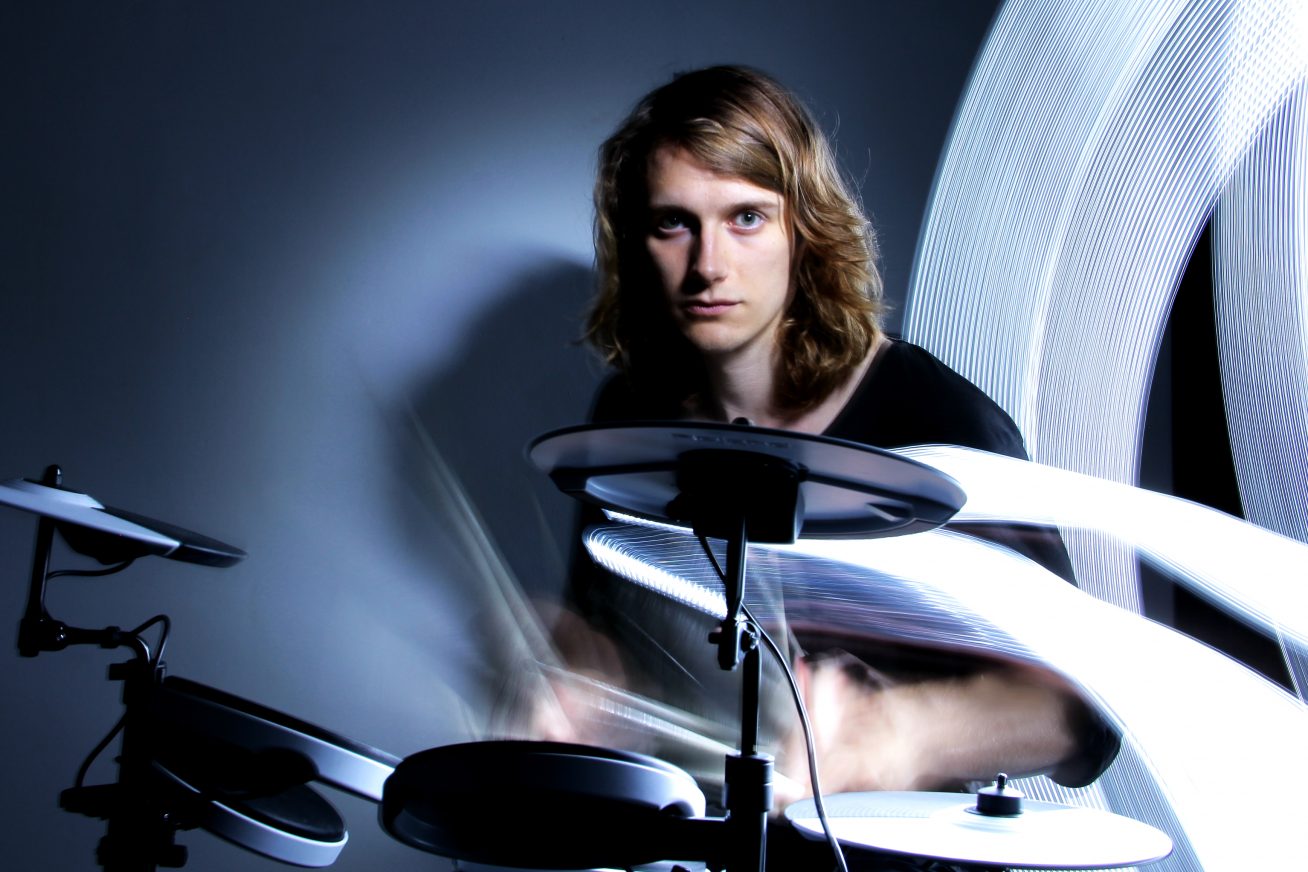 A young man will long, floppy blonde hair sits in a black t-shirt to the right of the picture. He swings drum sticks. In front of him is a high-tech drum kit. behind is a grey wall.