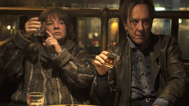 In a run down looking pub, two disheveled prople sit at a bar. On the left is a woman who holds up her tumbler, and points at it as though asking for a refill. On the right, a man with a long fringe also holds out his glass, at a slightly tipsy looking angle. This picture is from the film Can You Ever Forgive Me?