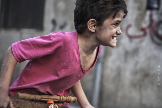 A young boy leans across this still from the film Capernaum. He wears a lose, v-neck pink shirt, and looks quite thin. His skin is covered in a sort of muddy dirt. He has dark hair, and his expression looks like a mix of desperation and pain.