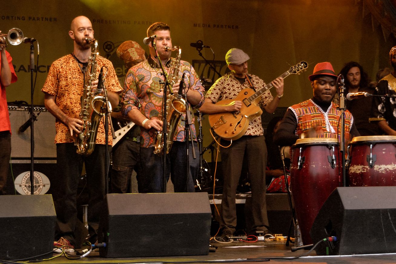 A band of musicians stand side by side on stage playing various instruments