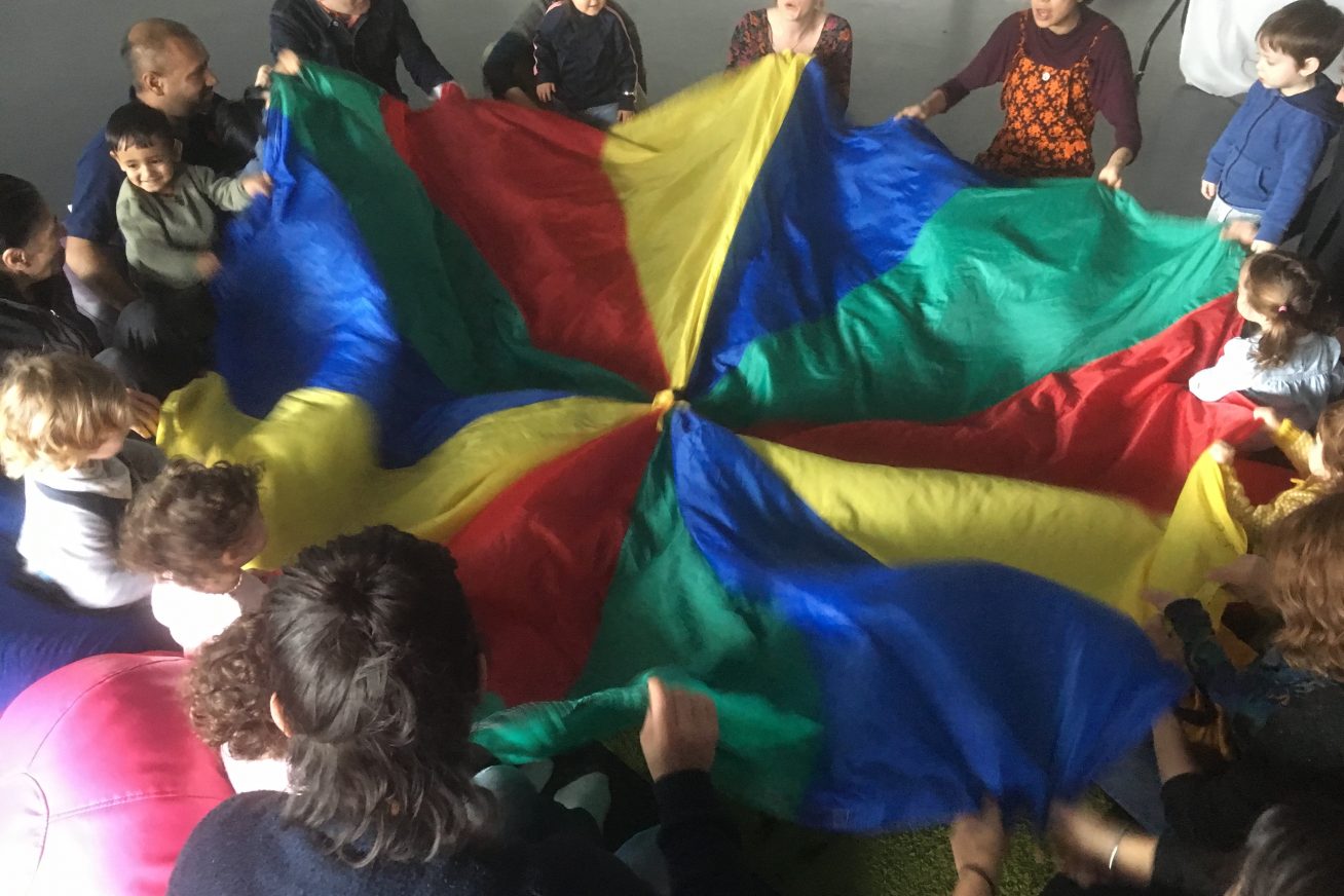 Children and adults play together with a rainbow coloured parachute