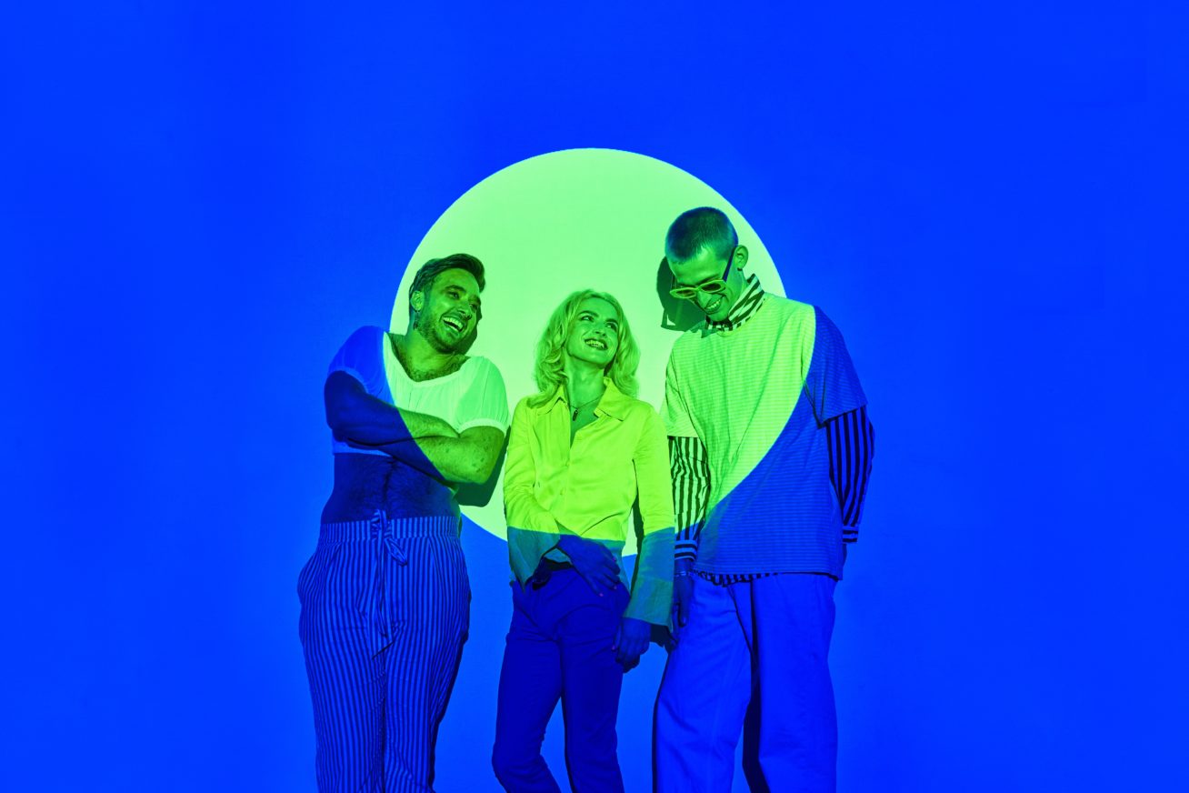 three people smiling in blue background, their faces highlighted by bright neon circle